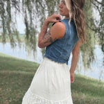 A woman outdoors by a lake wearing a sleeveless denim vest over a white lace skirt, photographed from the back with weeping willow branches hanging in the background.