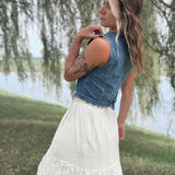 A woman outdoors by a lake wearing a sleeveless denim vest over a white lace skirt, photographed from the back with weeping willow branches hanging in the background.