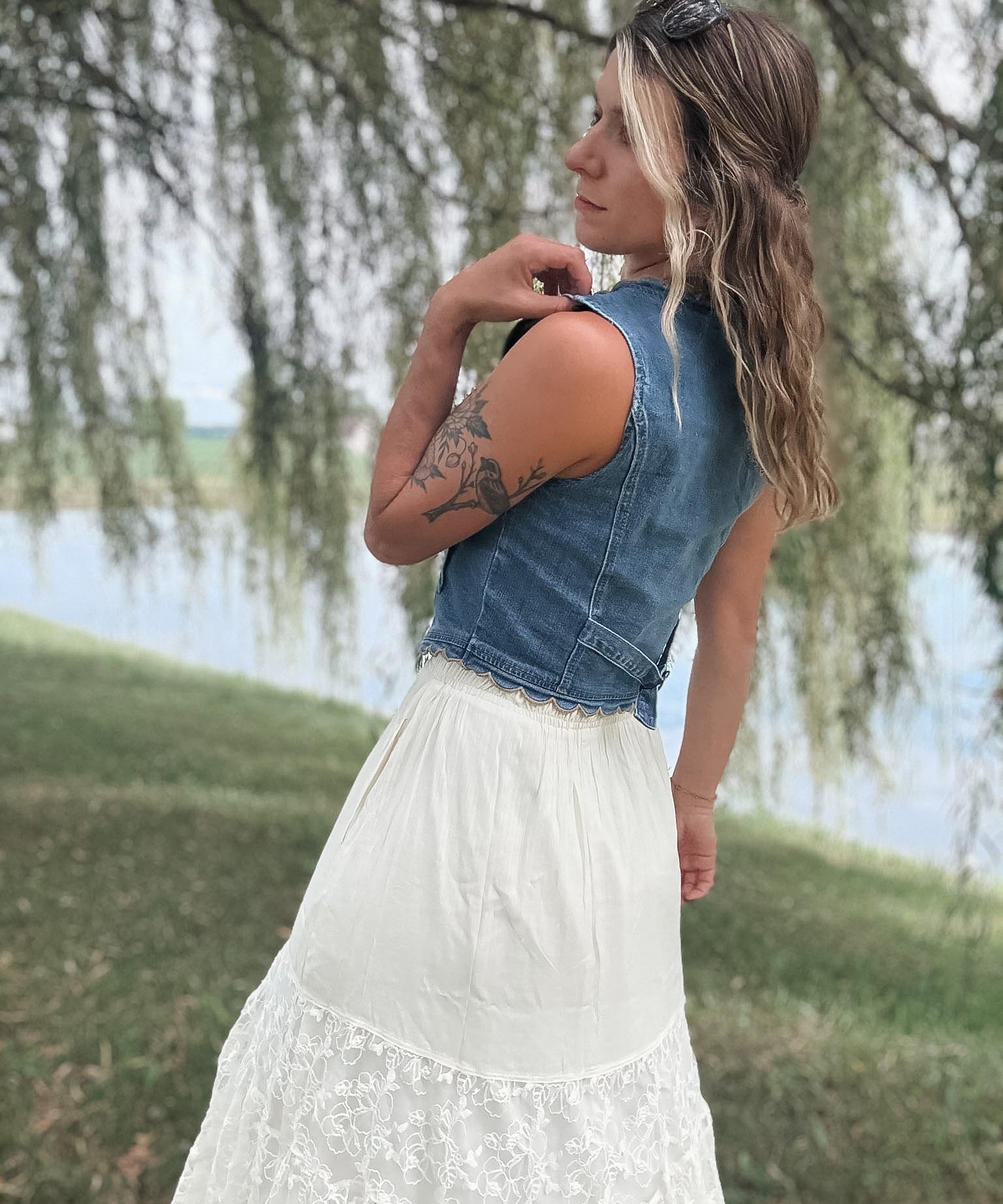 A woman outdoors by a lake wearing a sleeveless denim vest over a white lace skirt, photographed from the back with weeping willow branches hanging in the background.