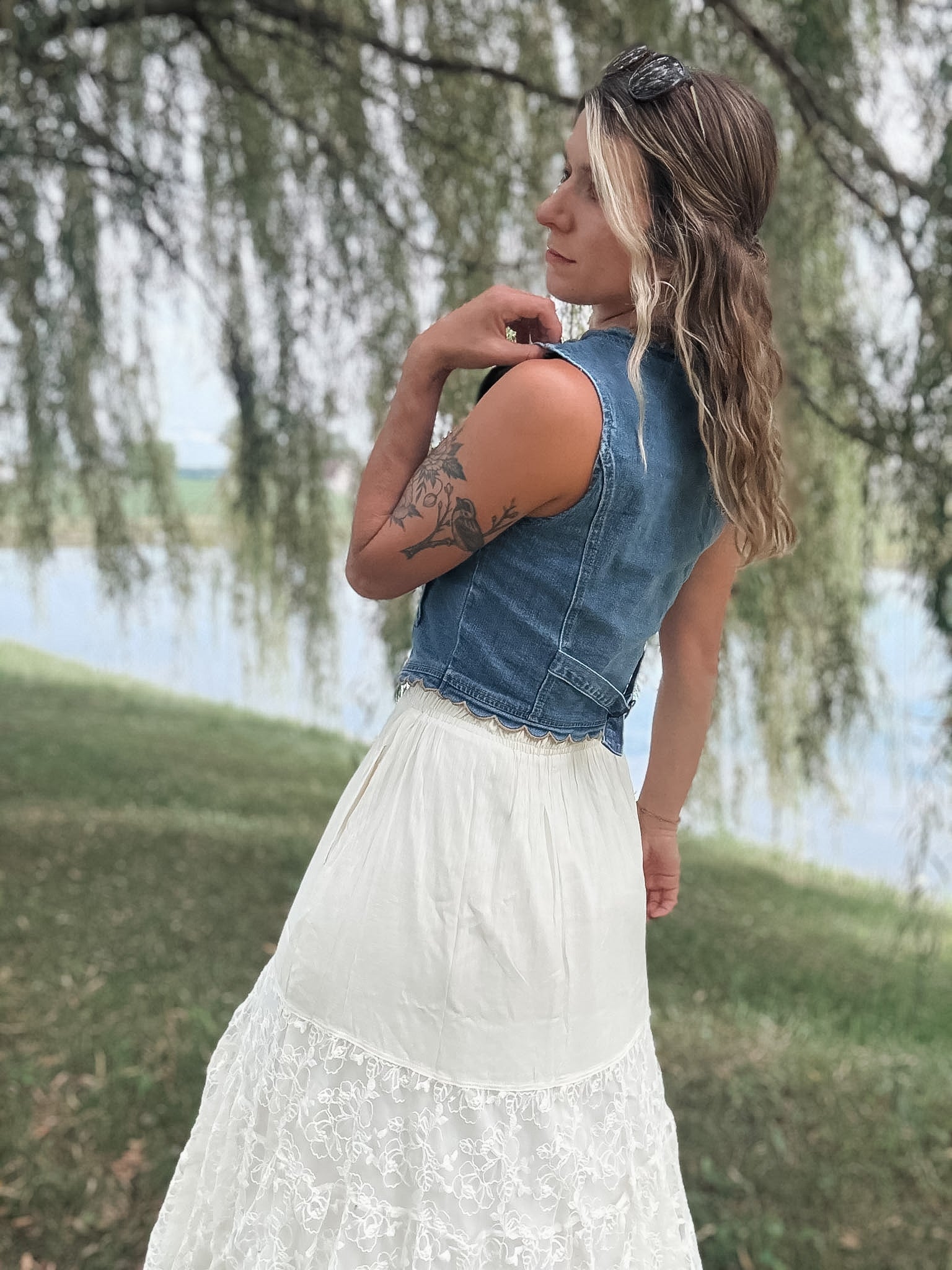 A woman outdoors by a lake wearing a sleeveless denim vest over a white lace skirt, photographed from the back with weeping willow branches hanging in the background.