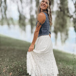 A woman wearing a sleeveless denim vest and a long white lace maxi skirt stands outdoors in a grassy area near water, holding out the skirt.