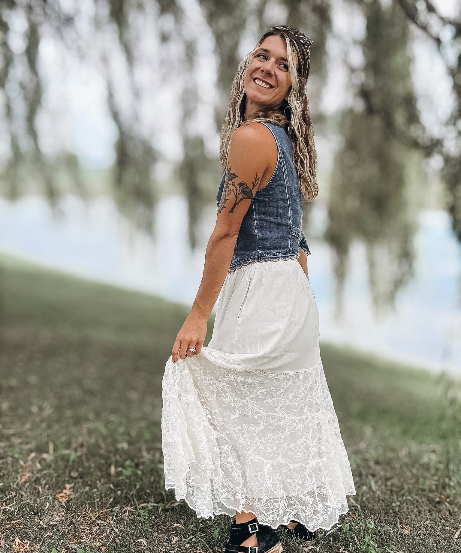 A woman wearing a sleeveless denim vest and a long white lace maxi skirt stands outdoors in a grassy area near water, holding out the skirt.