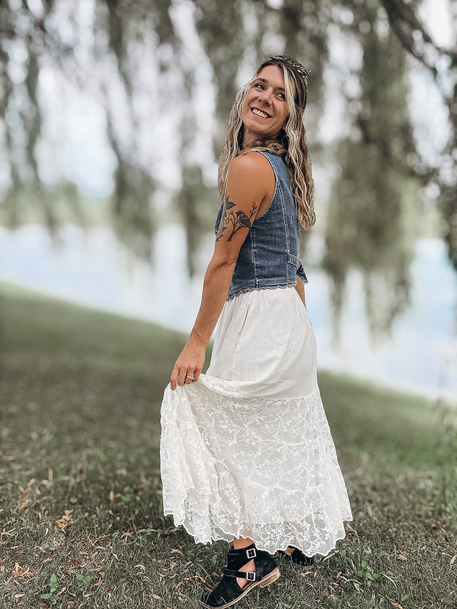 A woman wearing a sleeveless denim vest and a long white lace maxi skirt stands outdoors in a grassy area near water, holding out the skirt.