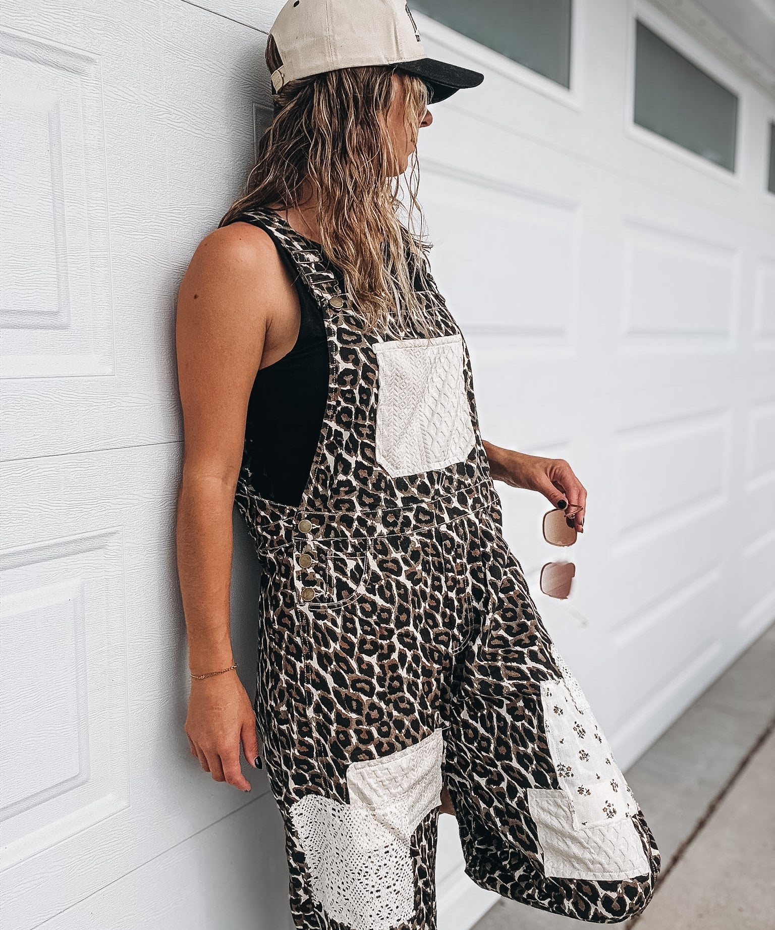 A woman wearing a black and brown leopard print overall jumpsuit with white patch pockets, a beige baseball cap, and sunglasses, leaning against a white garage door.