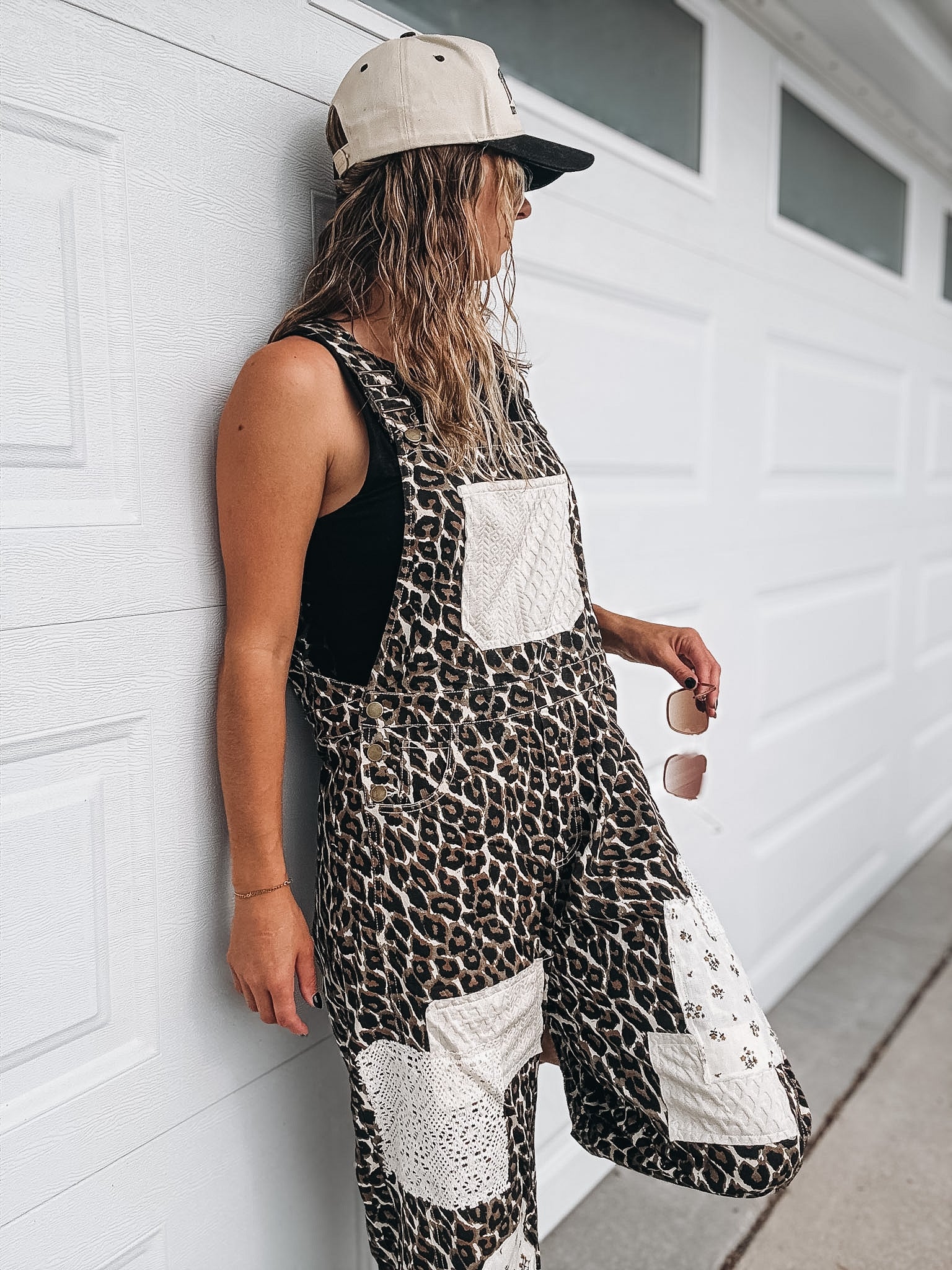 A woman wearing a black and brown leopard print overall jumpsuit with white patch pockets, a beige baseball cap, and sunglasses, leaning against a white garage door.