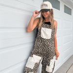 A woman wearing a leopard print overall jumpsuit with white patch pockets and a beige baseball cap stands against a white garage door.