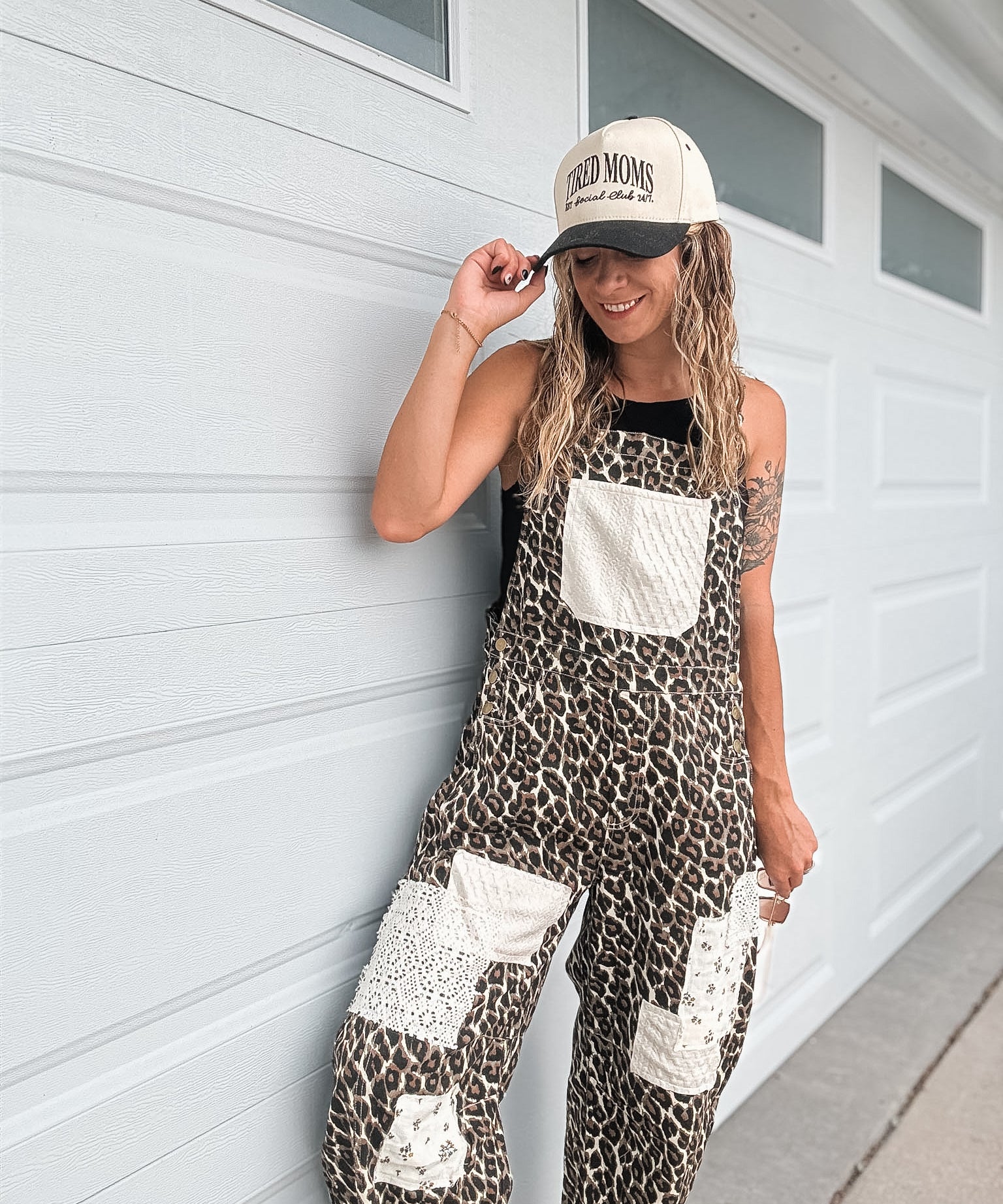 A woman wearing a leopard print overall jumpsuit with white patch pockets and a beige baseball cap stands against a white garage door.