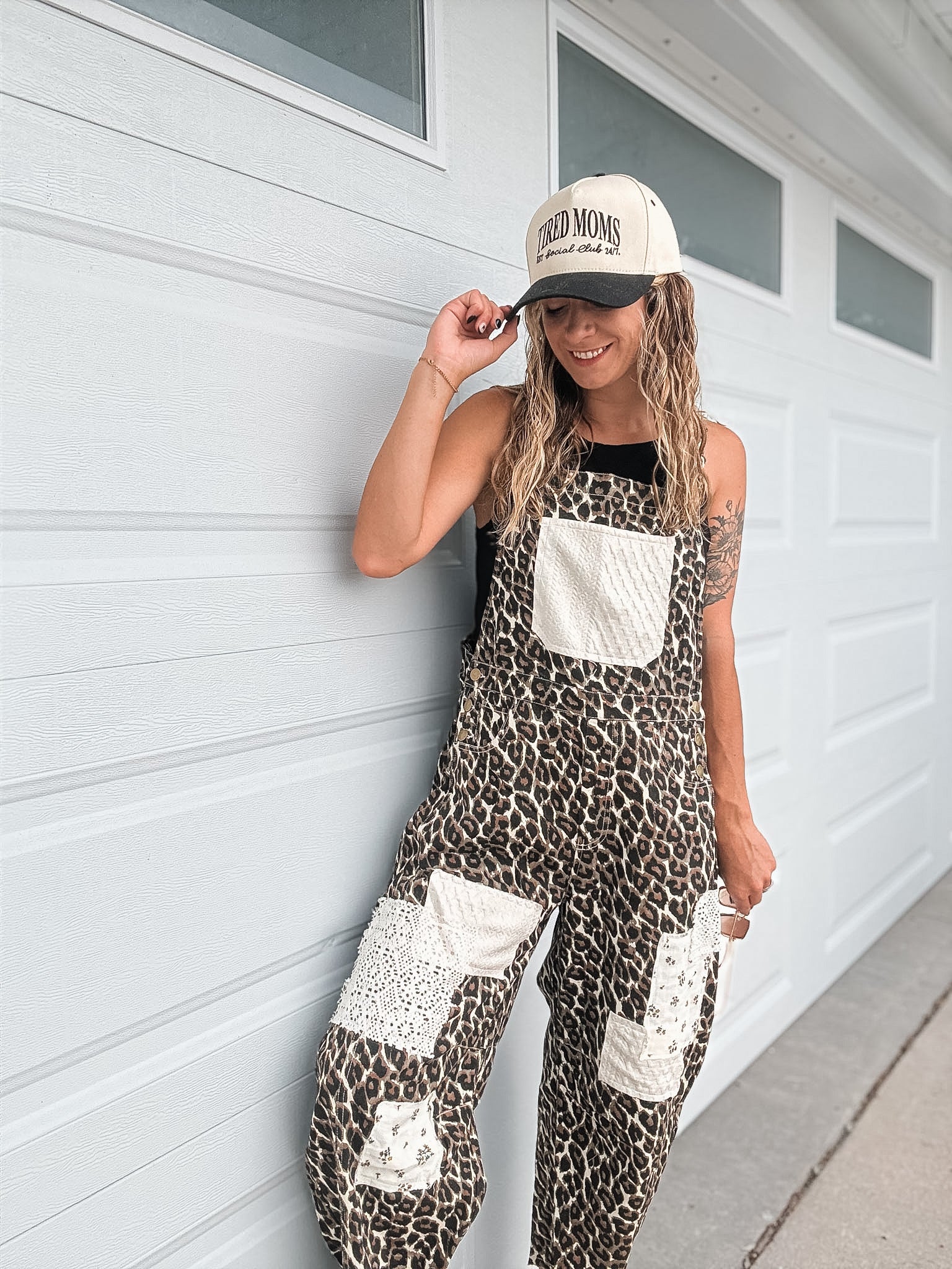A woman wearing a leopard print overall jumpsuit with white patch pockets and a beige baseball cap stands against a white garage door.