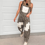 A woman wearing a sleeveless leopard-print overall jumpsuit with white patch pockets, a white baseball cap, and white sneakers, standing in front of a white garage door.