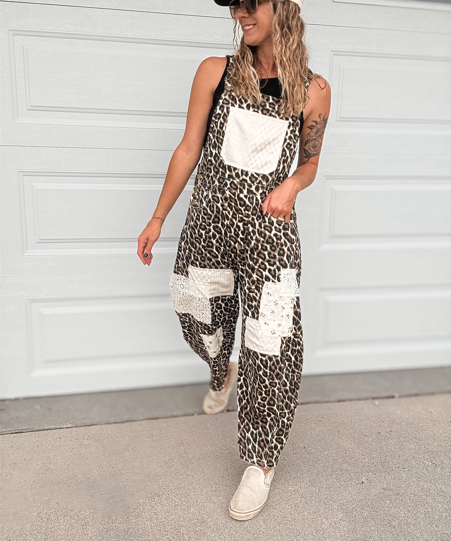 A woman wearing a sleeveless leopard-print overall jumpsuit with white patch pockets, a white baseball cap, and white sneakers, standing in front of a white garage door.