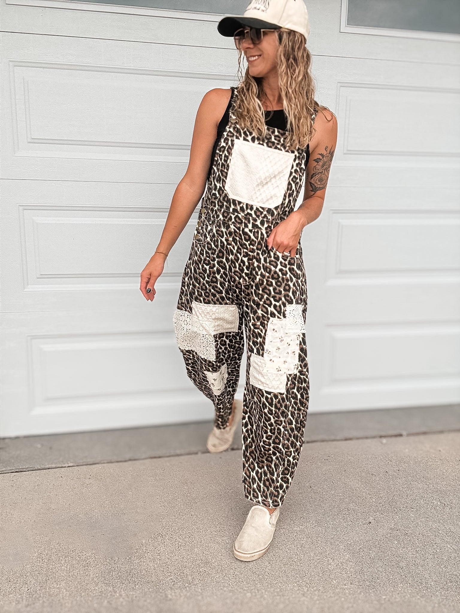 A woman wearing a sleeveless leopard-print overall jumpsuit with white patch pockets, a white baseball cap, and white sneakers, standing in front of a white garage door.