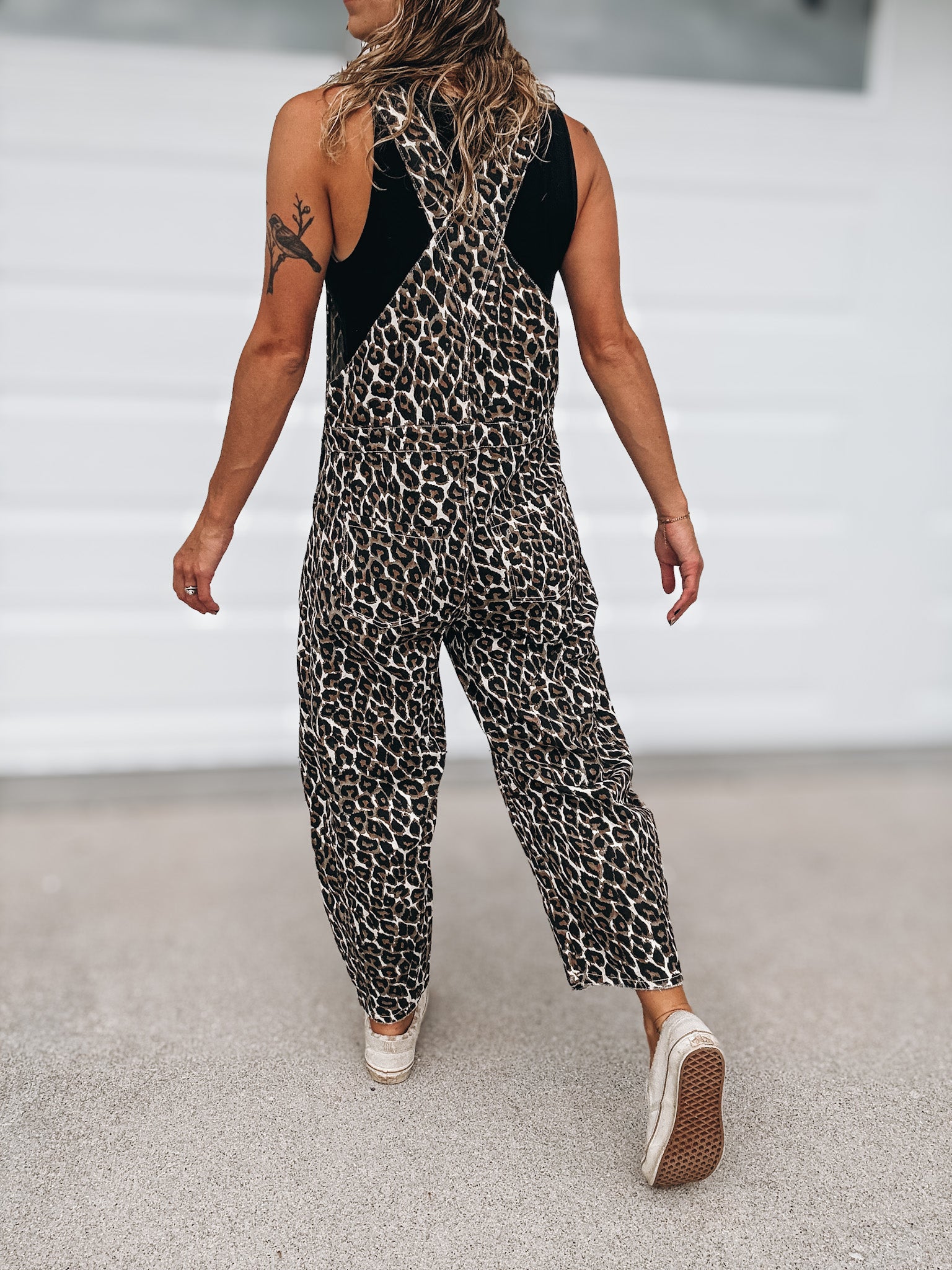 A woman wearing a sleeveless leopard-print jumpsuit with black shoulder panels, shown from behind as she walks on a light gray concrete surface.