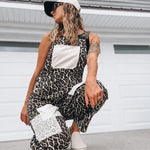 A woman crouches in front of a white garage door wearing a black and beige leopard-print overall jumpsuit with white patchwork pockets, a white baseball cap, and white slip-on shoes.