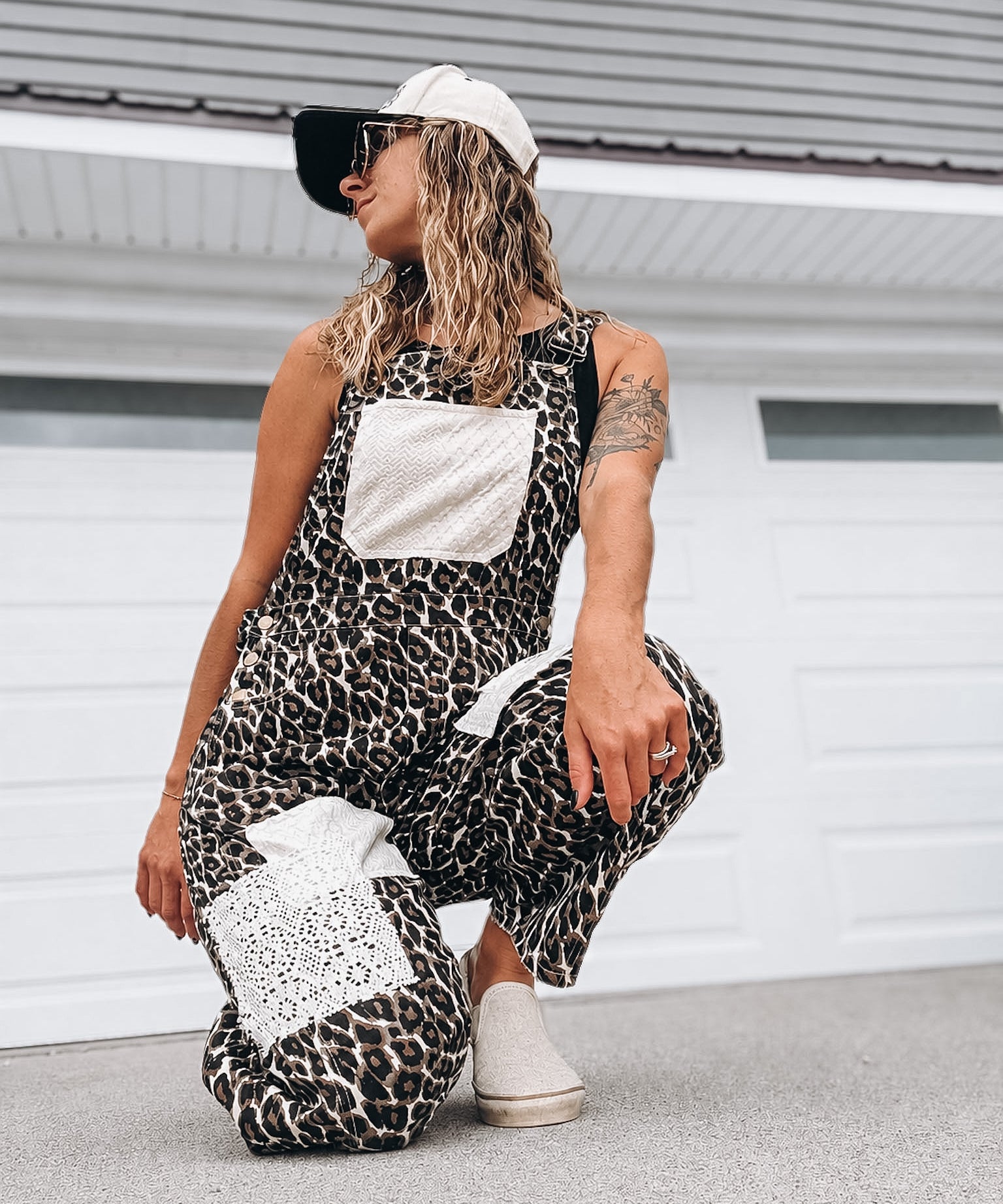 A woman crouches in front of a white garage door wearing a black and beige leopard-print overall jumpsuit with white patchwork pockets, a white baseball cap, and white slip-on shoes.
