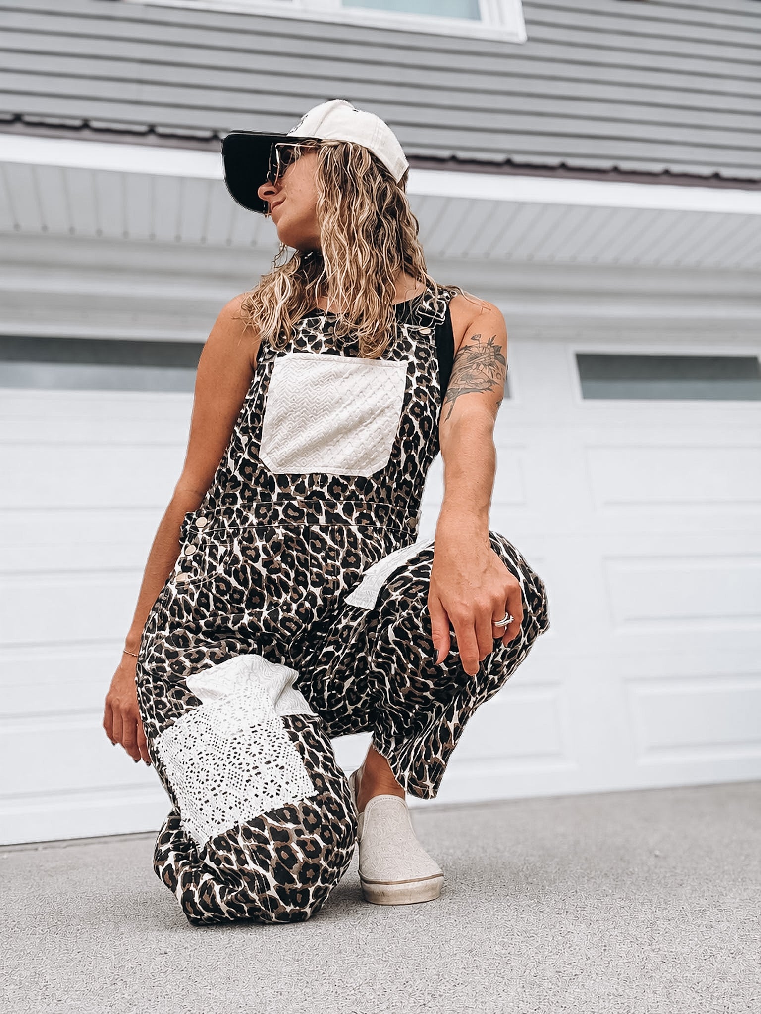 A woman crouches in front of a white garage door wearing a black and beige leopard-print overall jumpsuit with white patchwork pockets, a white baseball cap, and white slip-on shoes.