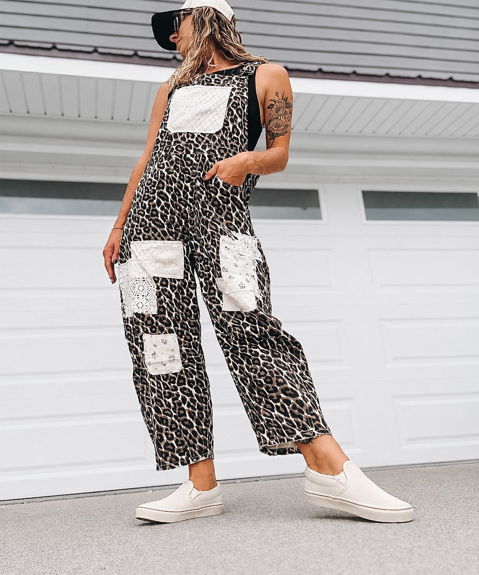 A woman wearing a black and white leopard-print sleeveless jumpsuit with white patch pockets, styled with a white cap, black visor, and white slip-on sneakers, standing in front of a white garage door.