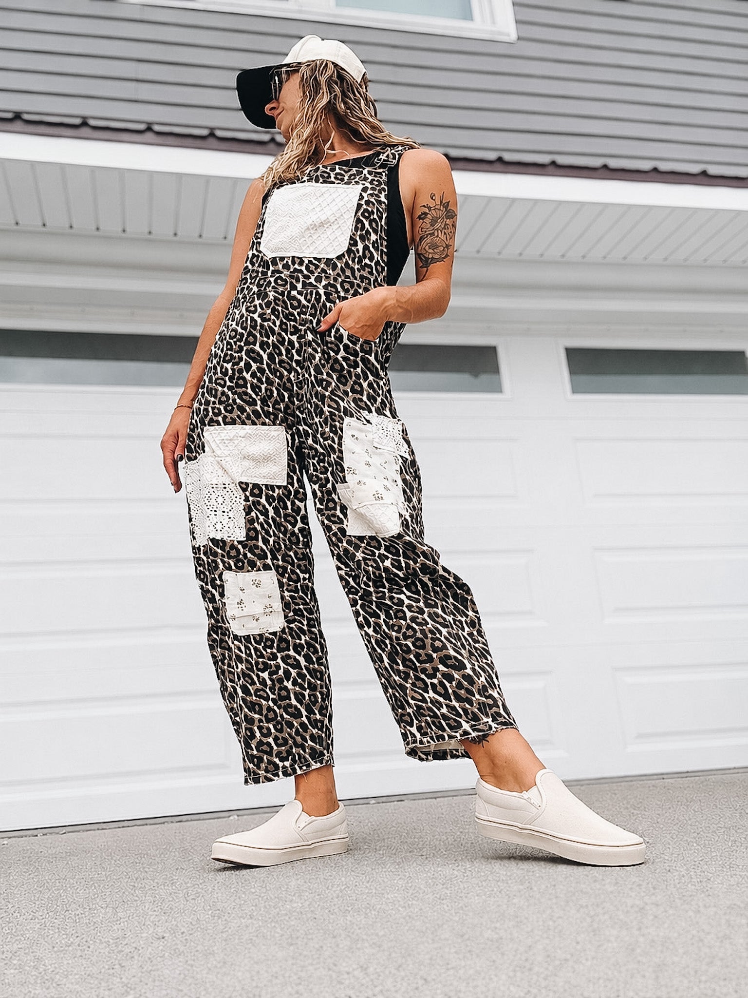 A woman wearing a black and white leopard-print sleeveless jumpsuit with white patch pockets, styled with a white cap, black visor, and white slip-on sneakers, standing in front of a white garage door.