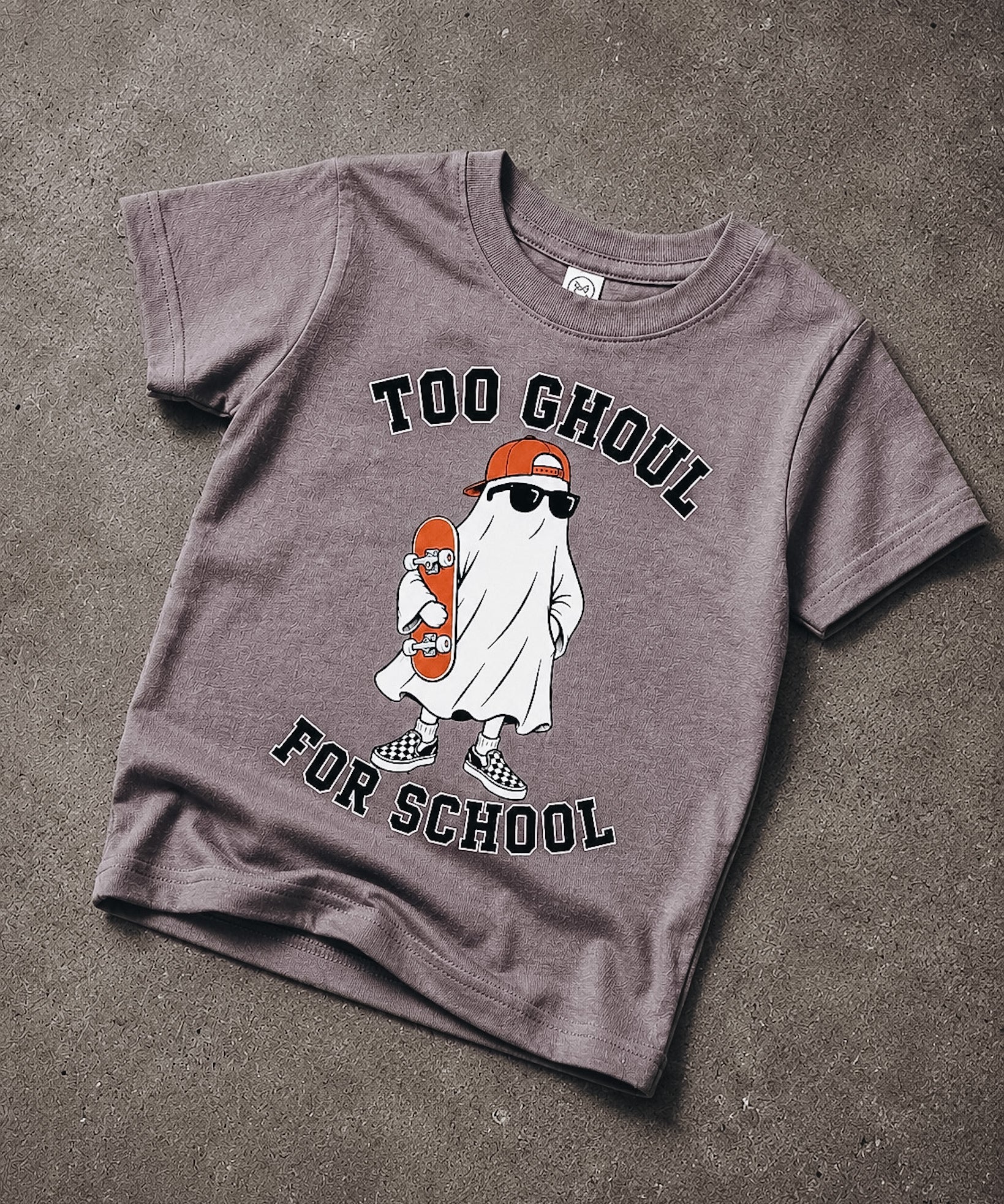 A short-sleeve crewneck t-shirt in a washed, heathered dark gray or charcoal color, laid flat on a textured concrete surface. The shirt features a graphic of a cartoon ghost wearing an orange cap and sunglasses while holding an orange skateboard, with the text 'TOO GHOUL FOR SCHOOL' printed in black arched above and below the illustration.