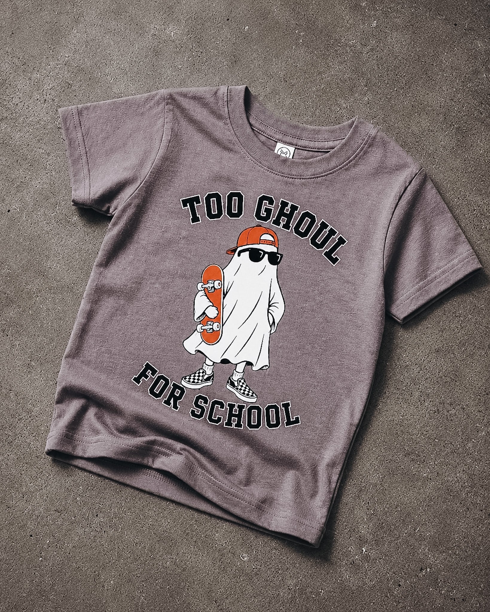 A short-sleeve crewneck t-shirt in a washed, heathered dark gray or charcoal color, laid flat on a textured concrete surface. The shirt features a graphic of a cartoon ghost wearing an orange cap and sunglasses while holding an orange skateboard, with the text 'TOO GHOUL FOR SCHOOL' printed in black arched above and below the illustration.