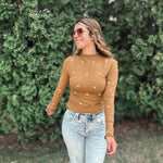 A woman wearing a fitted long-sleeve tan top with small white embroidered star details, paired with light-wash ripped jeans, standing outdoors in front of a green hedge.