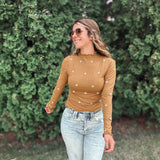 A woman wearing a fitted long-sleeve tan top with small white embroidered star details, paired with light-wash ripped jeans, standing outdoors in front of a green hedge.