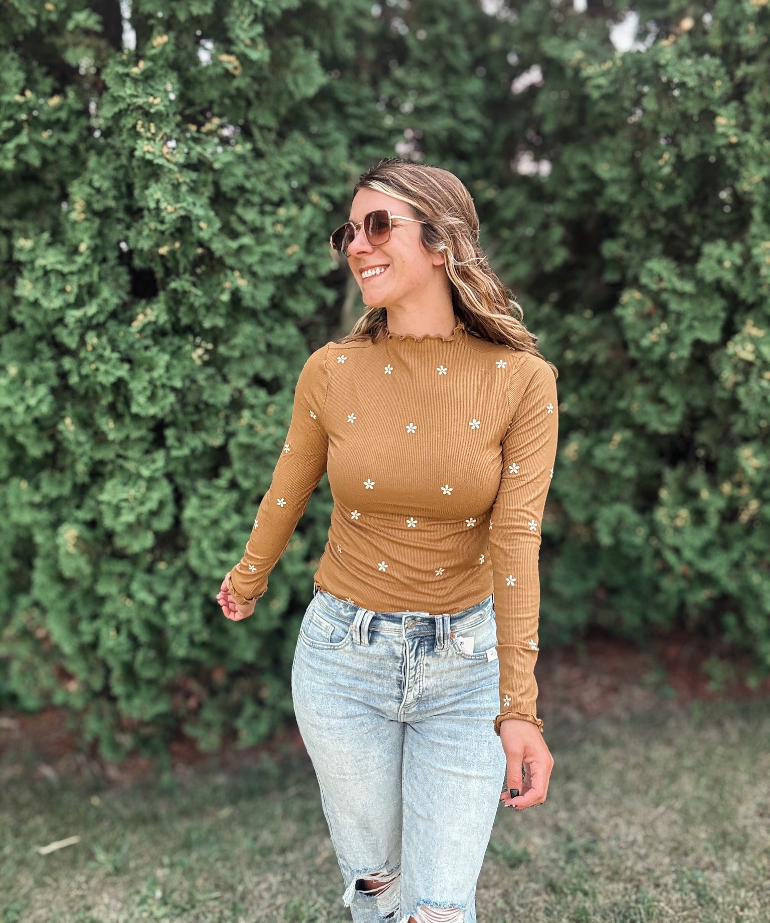 A woman wearing a fitted long-sleeve tan top with small white embroidered star details, paired with light-wash ripped jeans, standing outdoors in front of a green hedge.