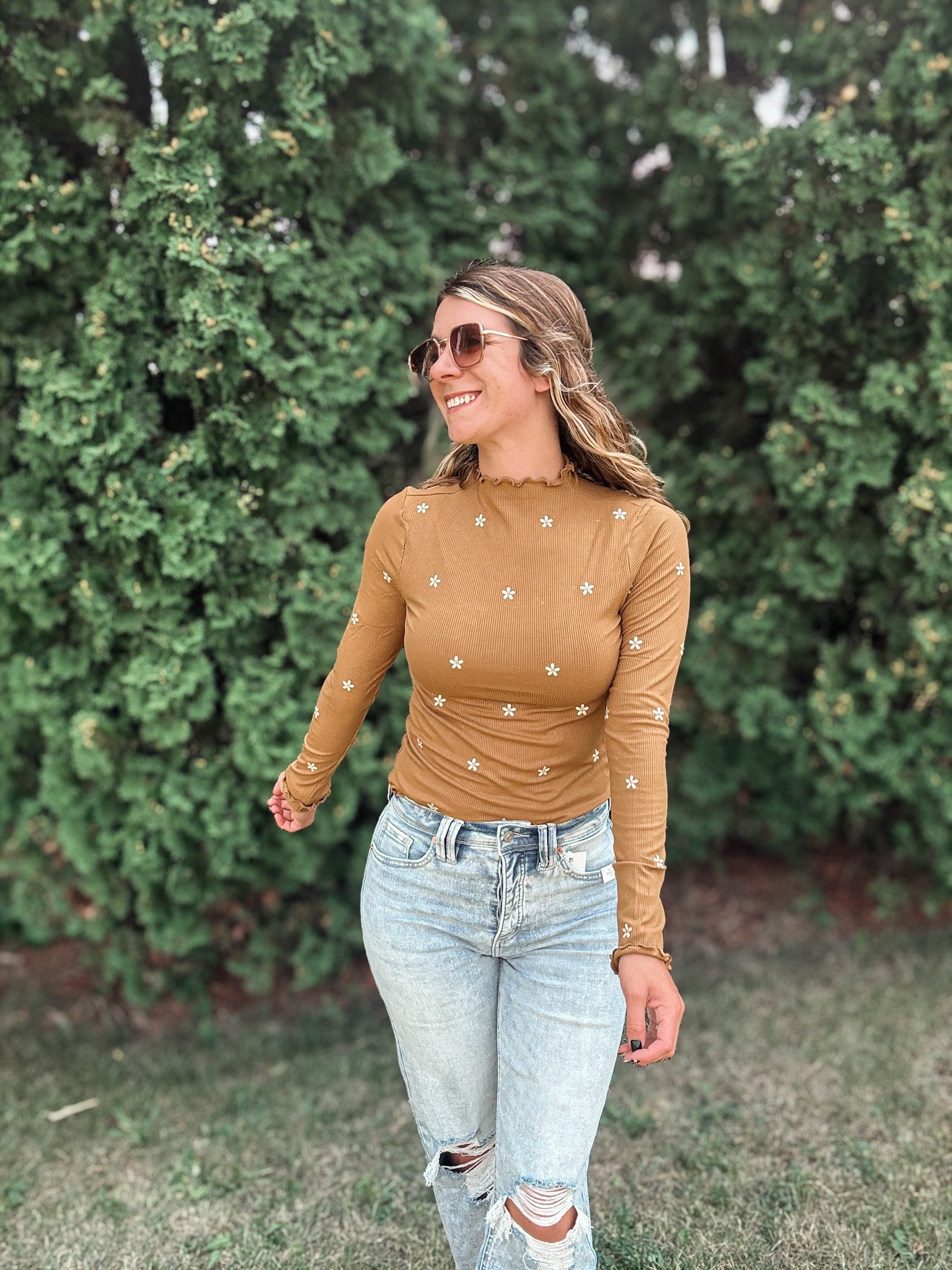 A woman wearing a fitted long-sleeve tan top with small white embroidered star details, paired with light-wash ripped jeans, standing outdoors in front of a green hedge.