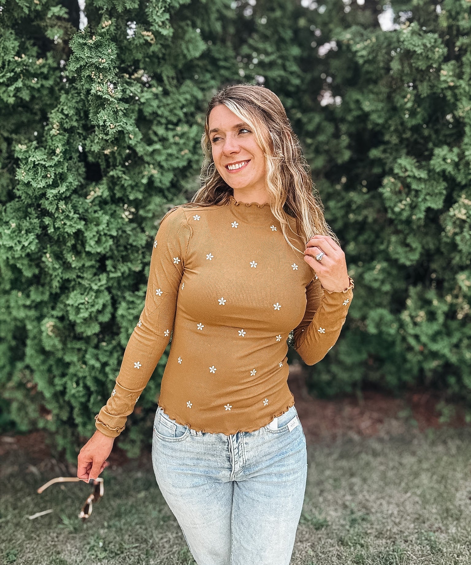 A woman wearing a fitted, long-sleeve tan top with small white embroidered star details, paired with light-wash blue jeans, standing outdoors in front of a green hedge.
