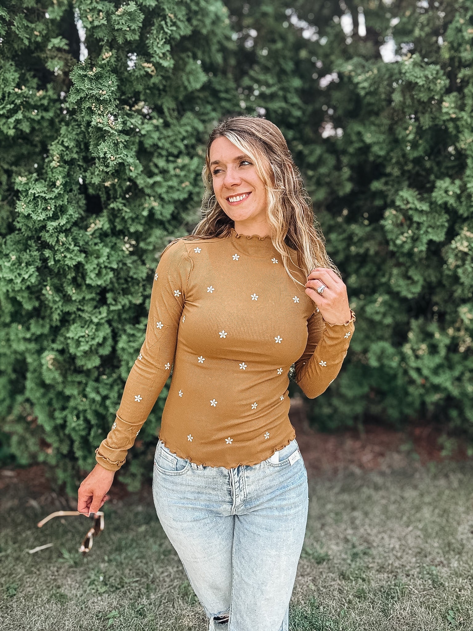 A woman wearing a fitted, long-sleeve tan top with small white embroidered star details, paired with light-wash blue jeans, standing outdoors in front of a green hedge.