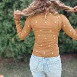 A woman viewed from behind wearing a tan long-sleeve top with small white embroidered flowers, paired with light blue jeans, set against a blurred green garden background.
