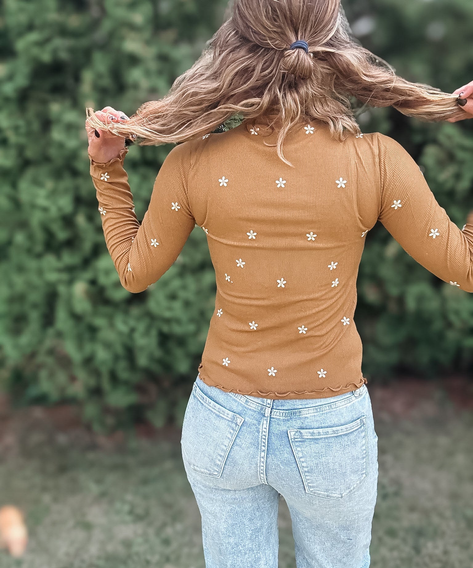 A woman viewed from behind wearing a tan long-sleeve top with small white embroidered flowers, paired with light blue jeans, set against a blurred green garden background.