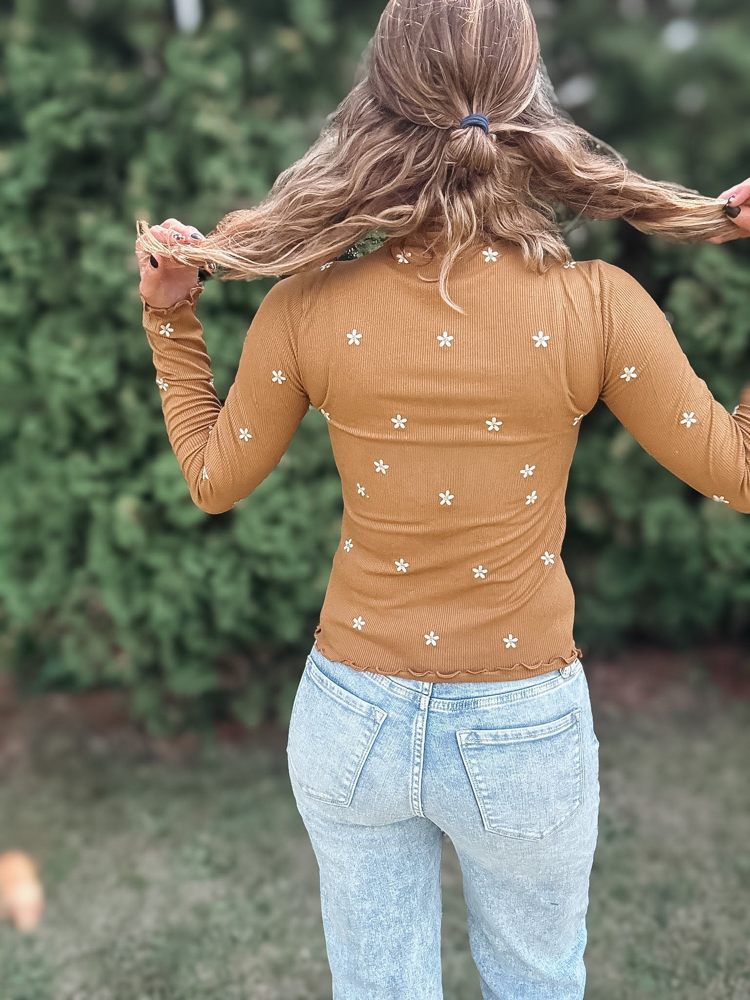 A woman viewed from behind wearing a tan long-sleeve top with small white embroidered flowers, paired with light blue jeans, set against a blurred green garden background.