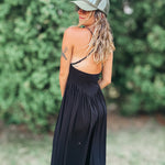 A woman wearing a black maxi dress with a backless open design, a green trucker hat, and sunglasses, photographed outdoors against a blurred green foliage background.