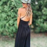 A woman wearing a black maxi dress with a backless open design, a green trucker hat, and sunglasses, photographed outdoors against a blurred green foliage background.