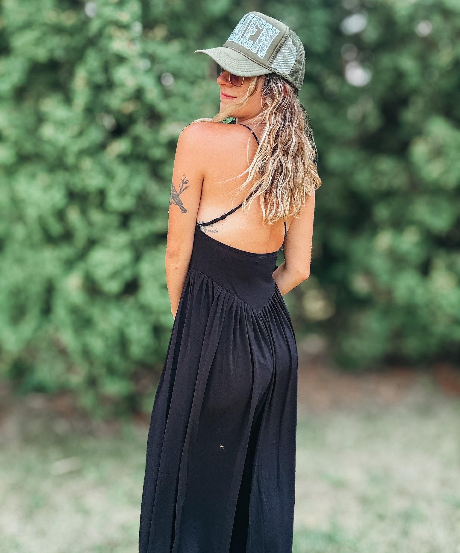 A woman wearing a black maxi dress with a backless open design, a green trucker hat, and sunglasses, photographed outdoors against a blurred green foliage background.