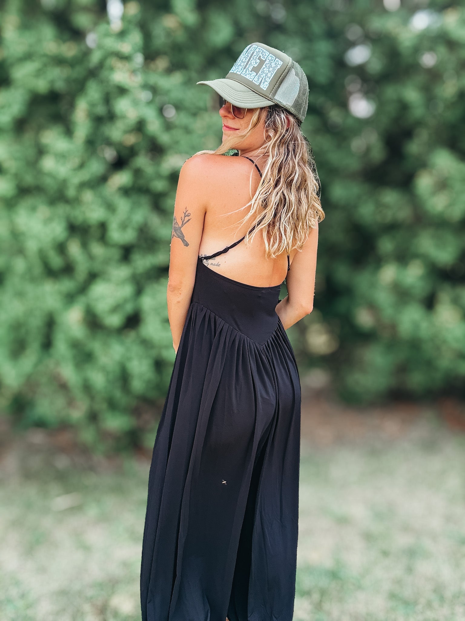 A woman wearing a black maxi dress with a backless open design, a green trucker hat, and sunglasses, photographed outdoors against a blurred green foliage background.