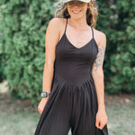 A woman wearing a black sleeveless jumpsuit with thin straps and a gathered waist, styled with a camouflage baseball cap that says 'AMEN' in glittery letters, standing outdoors in front of a green hedge.
