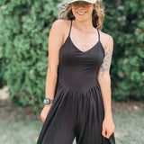 A woman wearing a black sleeveless jumpsuit with thin straps and a gathered waist, styled with a camouflage baseball cap that says 'AMEN' in glittery letters, standing outdoors in front of a green hedge.
