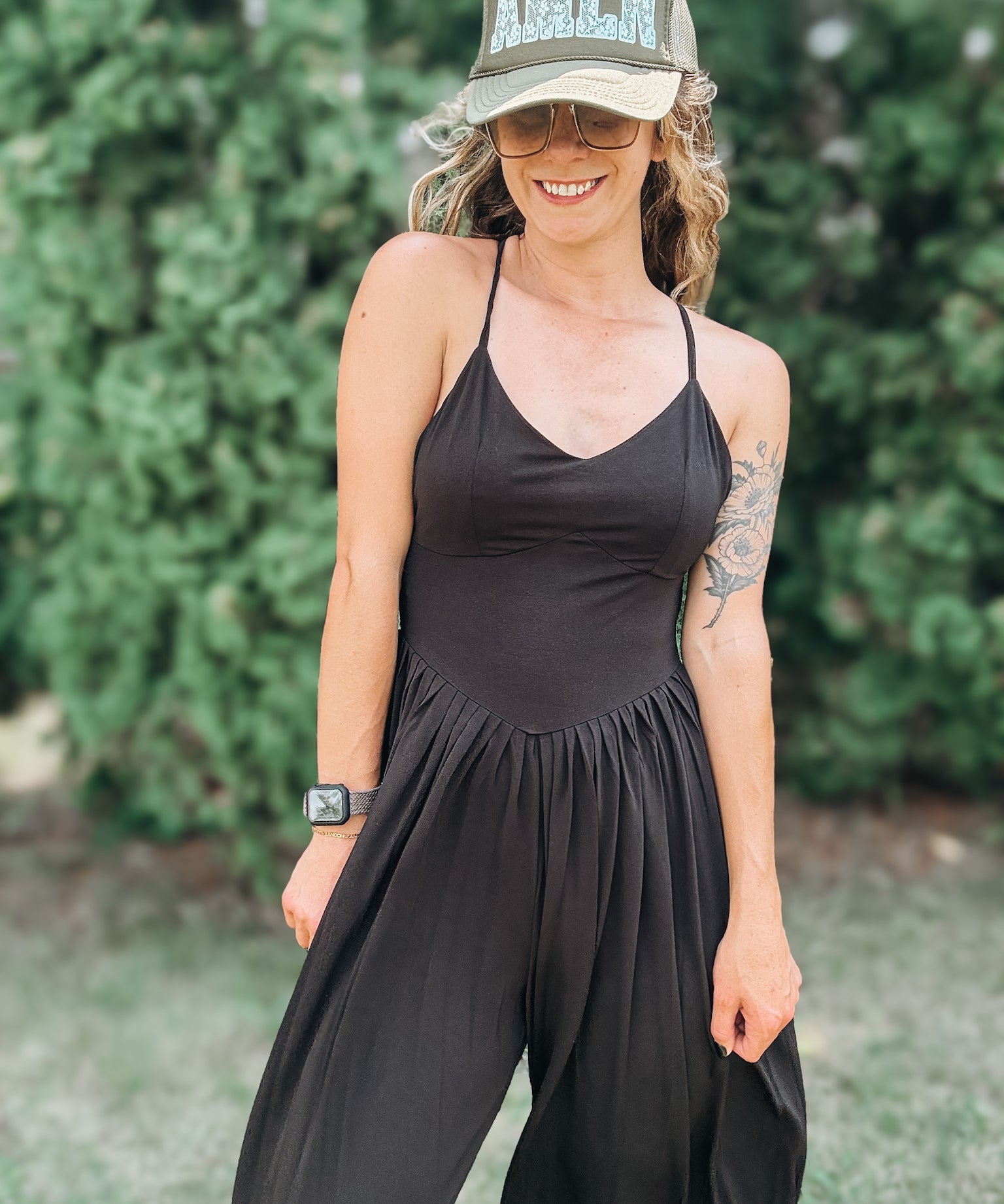A woman wearing a black sleeveless jumpsuit with thin straps and a gathered waist, styled with a camouflage baseball cap that says 'AMEN' in glittery letters, standing outdoors in front of a green hedge.