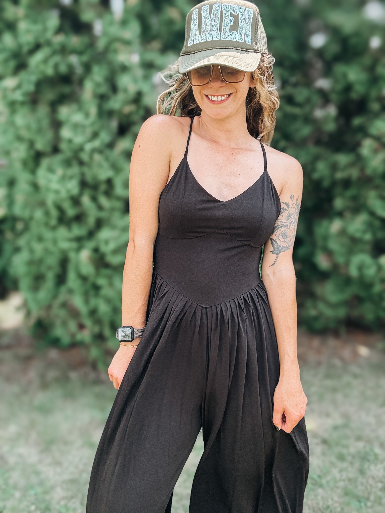 A woman wearing a black sleeveless jumpsuit with thin straps and a gathered waist, styled with a camouflage baseball cap that says 'AMEN' in glittery letters, standing outdoors in front of a green hedge.
