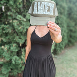 A woman wearing a black dress holds a green trucker hat in front of her face, which displays the word 'AMEN' in a textured white font. The background is a soft-focus outdoor setting with green foliage.