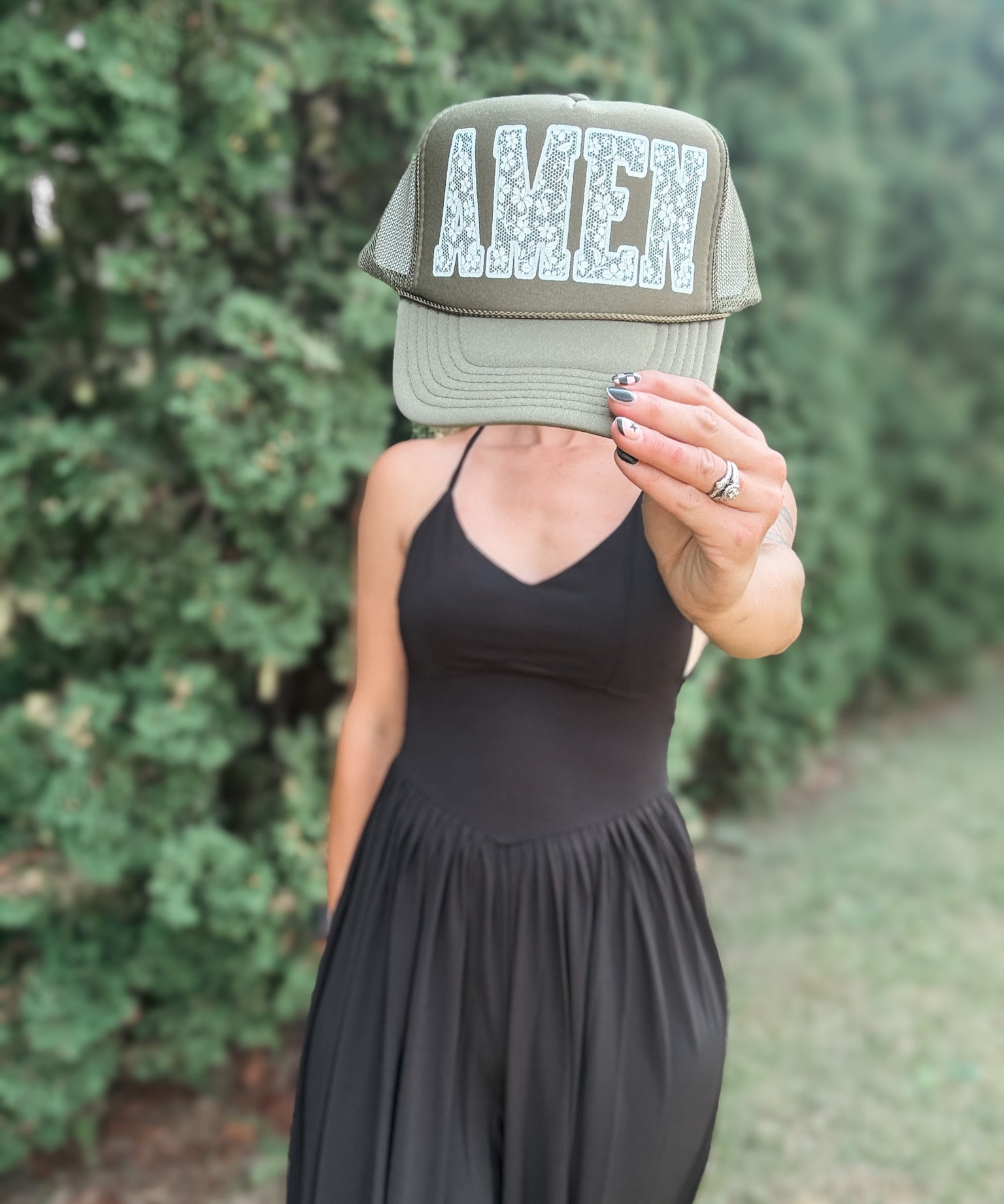 A woman wearing a black dress holds a green trucker hat in front of her face, which displays the word 'AMEN' in a textured white font. The background is a soft-focus outdoor setting with green foliage.