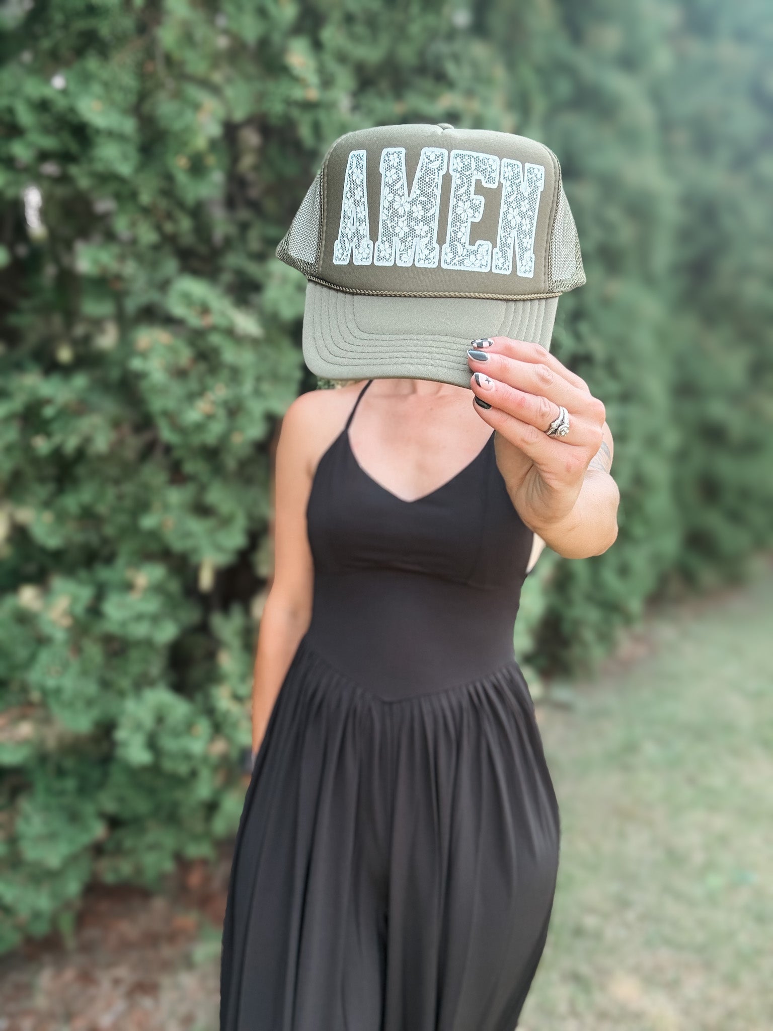 A woman wearing a black dress holds a green trucker hat in front of her face, which displays the word 'AMEN' in a textured white font. The background is a soft-focus outdoor setting with green foliage.