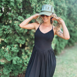 A woman wearing a black sleeveless maxi dress and a green trucker hat with 'AMEN' in lace letters, posing outdoors in front of a green hedge.