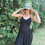 A woman wearing a black sleeveless maxi dress and a green trucker hat with 'AMEN' in lace letters, posing outdoors in front of a green hedge.