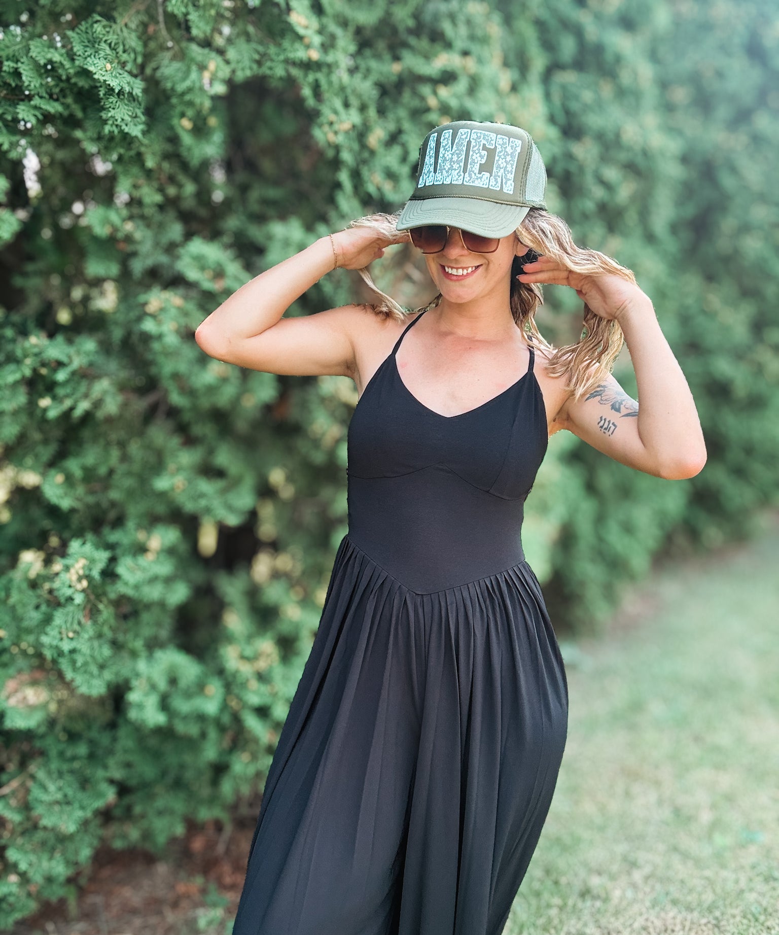 A woman wearing a black sleeveless maxi dress and a green trucker hat with 'AMEN' in lace letters, posing outdoors in front of a green hedge.