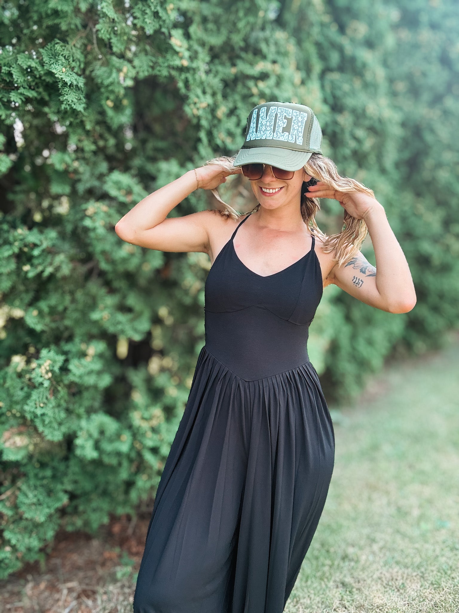 A woman wearing a black sleeveless maxi dress and a green trucker hat with 'AMEN' in lace letters, posing outdoors in front of a green hedge.