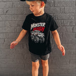 A young boy wearing a black t-shirt with a 'Monster Mash' monster truck graphic, gray shorts, and a black baseball cap, standing against a gray brick wall.
