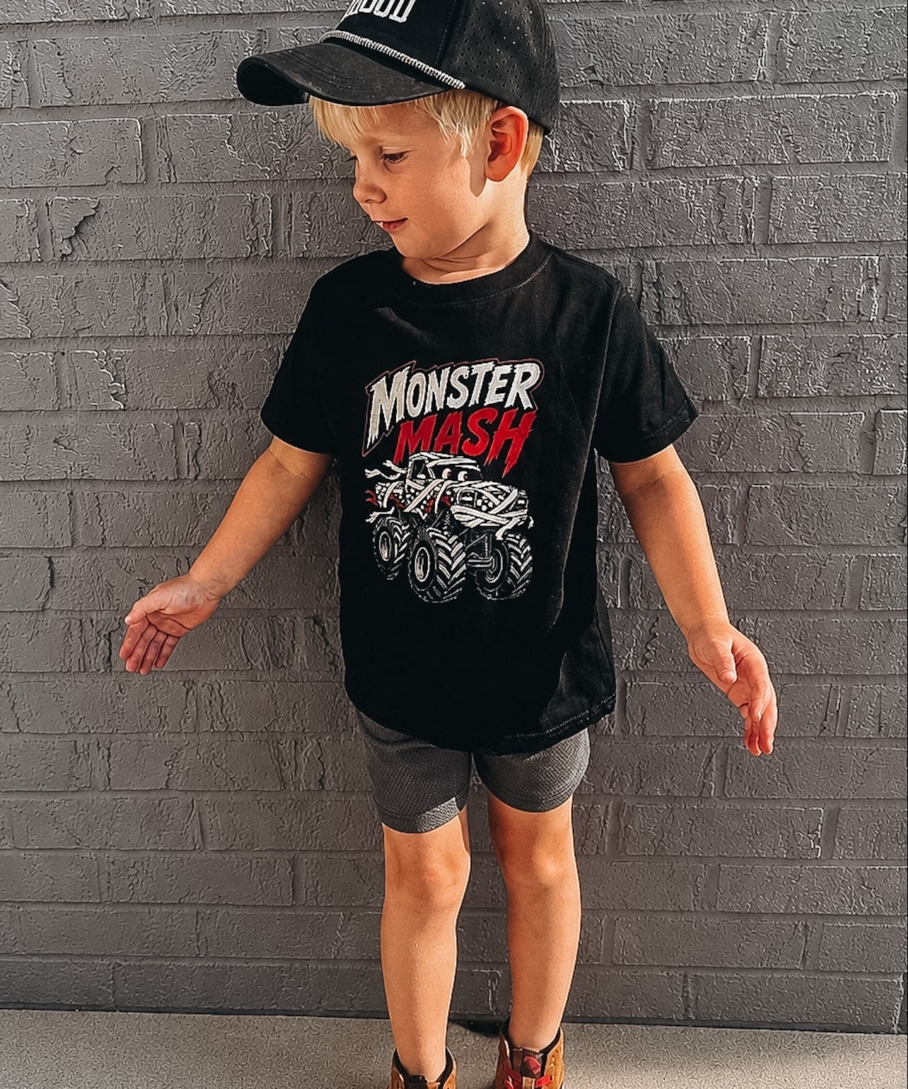 A young boy wearing a black t-shirt with a 'Monster Mash' monster truck graphic, gray shorts, and a black baseball cap, standing against a gray brick wall.