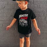 A young boy wearing a black t-shirt with a 'Monster Mash' monster truck graphic, gray shorts, and a black baseball cap, standing against a gray brick wall.